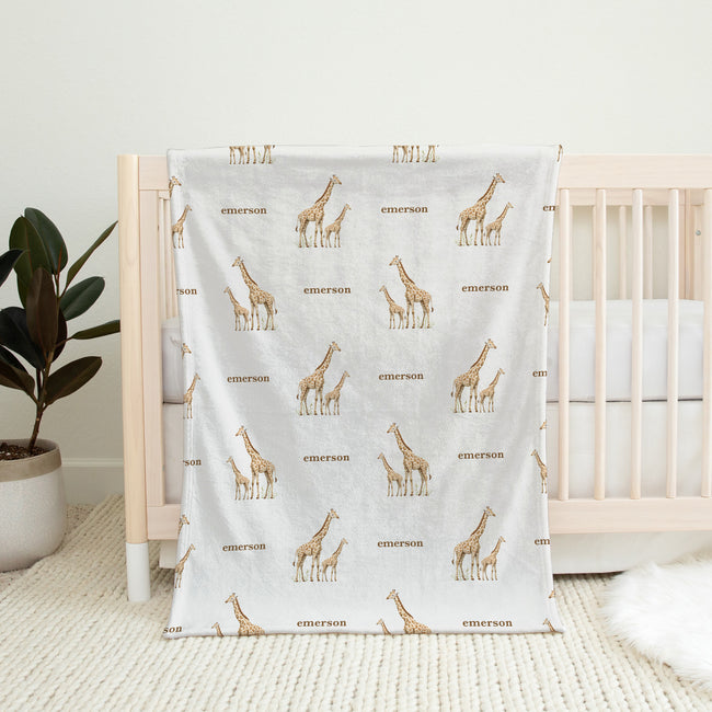 A baby blanket with a repeating pattern of brown giraffes and the name 'emerson' in brown text, draped over a light wood crib in a nursery setting.