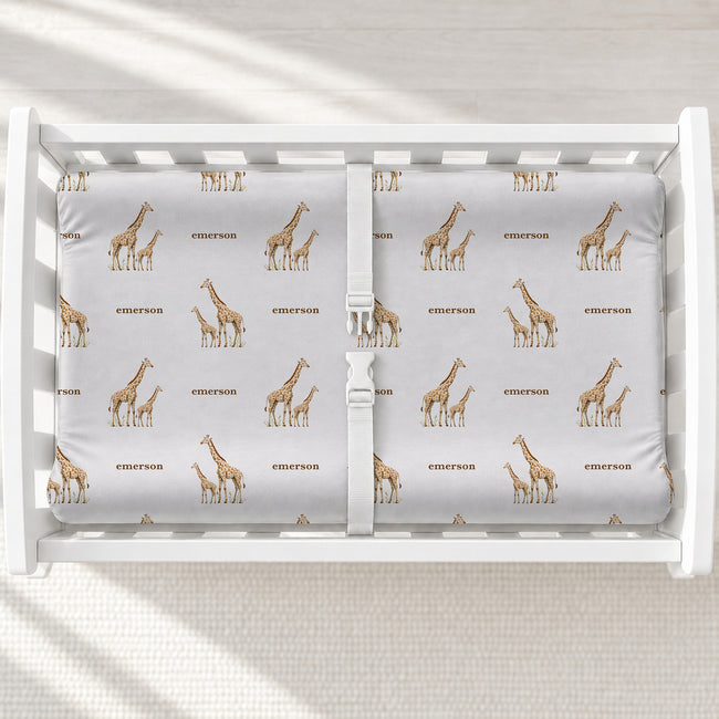 A changing table with a changing pad cover featuring a repeating giraffe and text pattern in brown and beige on a light gray background.