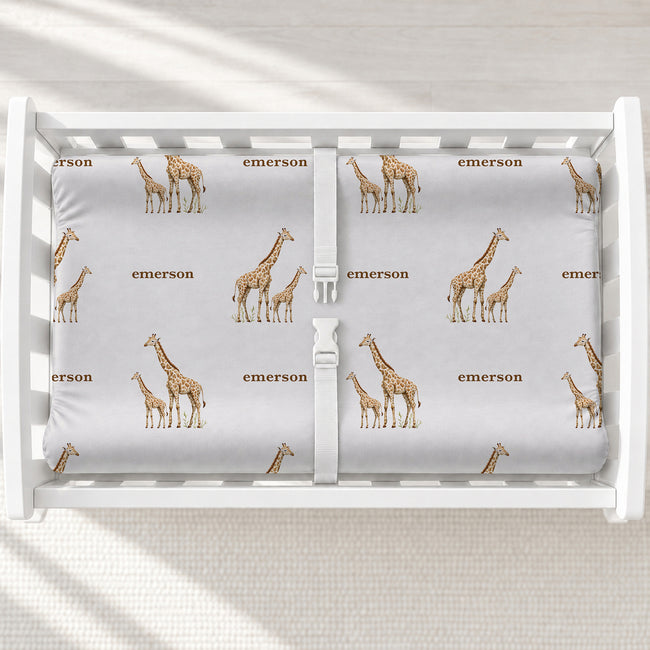 Little Giraffe personalized changing pad cover featuring watercolor adult and baby giraffes and the name Emerson in brown, shown on a white changing table.