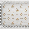 A white wooden crib with a fitted sheet featuring a repeating pattern of brown giraffes and the word 'Emerson' in brown text, shown from a top-down angle on a light textured floor.