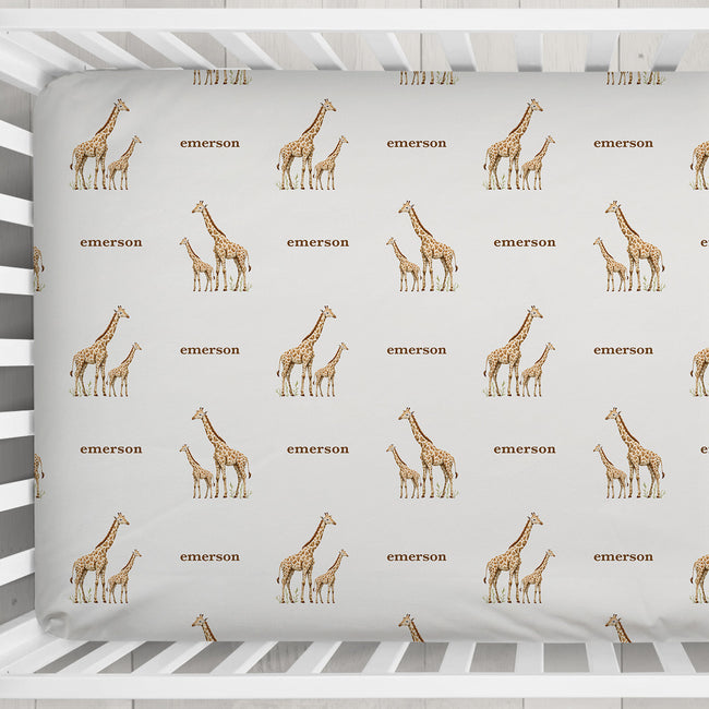 A white wooden crib with a fitted sheet featuring a repeating pattern of brown giraffes and the word 'Emerson' in brown text, shown from a top-down angle on a light textured floor.