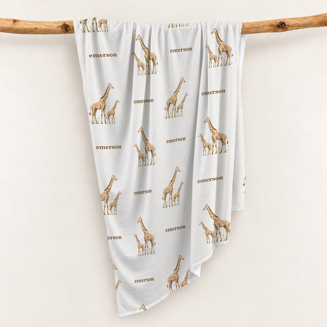Little Giraffe swaddle blanket personalized with Emerson, draped over a wooden branch to display the repeating giraffe pattern.