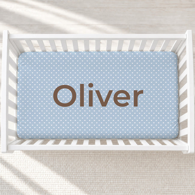 A light blue baby crib mattress cover with white polka dots and the name 'Oliver' printed in brown, shown inside a white wooden crib.