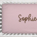 A pink baby crib mattress cover with a white heart pattern and the name 'Sophie' printed in brown script, shown inside a white wooden crib.