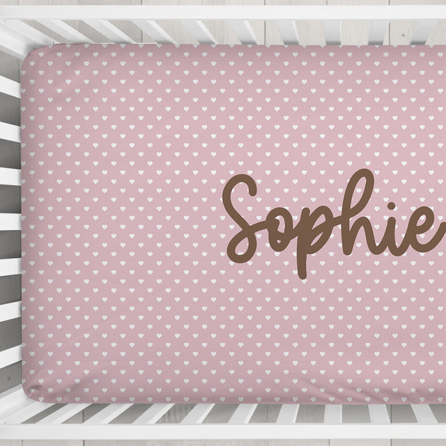 A pink baby crib mattress cover with a white heart pattern and the name 'Sophie' printed in brown script, shown inside a white wooden crib.