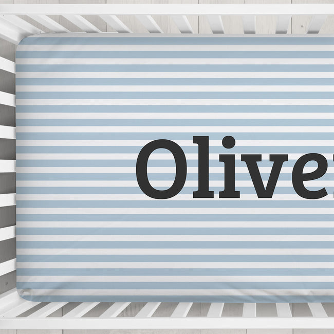 A personalized baby crib sheet with light blue and white horizontal stripes, featuring the name 'Oliver' printed in large black serif letters, shown inside a white wooden crib.
