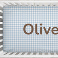 A light blue and white houndstooth-patterned fitted crib sheet with the name 'Oliver' printed in large brown letters, shown inside a white crib.