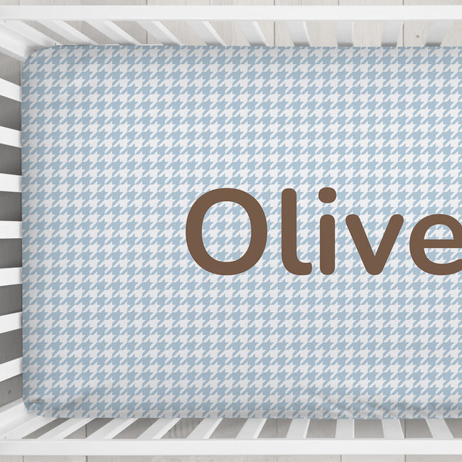 A light blue and white houndstooth-patterned fitted crib sheet with the name 'Oliver' printed in large brown letters, shown inside a white crib.