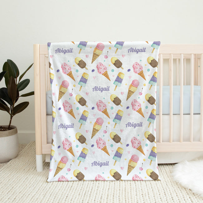 A personalized baby blanket with a colorful ice cream cone pattern and the name 'Abigail' printed repeatedly, draped over a light wood crib in a nursery setting.