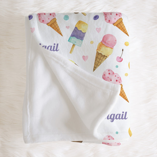 A white baby blanket with a colorful watercolor ice cream and popsicle print, featuring the name 'Abigail' in purple script, laid flat on a white faux fur background.