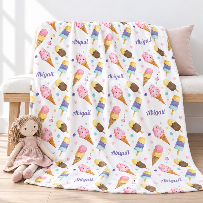 Personalized watercolor ice cream blanket with Abigail in purple script, draped over a light wood bench with a pink cloth doll resting on the edge.