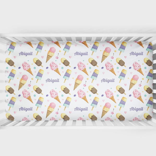 A crib bumper sticker or insert featuring a repeating pattern of colorful watercolor ice cream cones, cherries, hearts, and the name 'Abigail' in purple script, displayed inside a white wooden crib.
