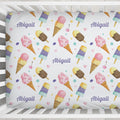 A baby bedding set featuring a custom printed sheet or bumper sticker with a watercolor ice cream cone pattern, pink and purple text reading 'Abigail', and small pink hearts on a white background, shown inside a white crib.