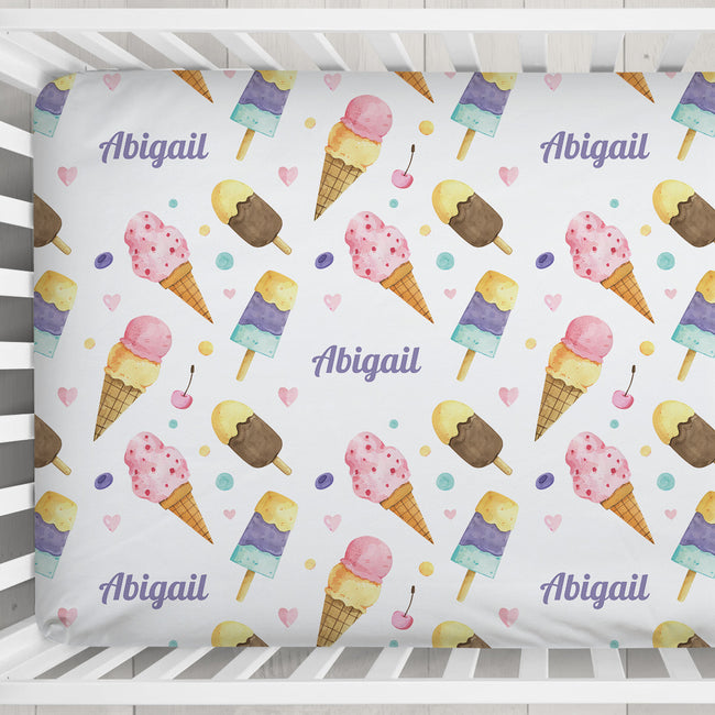 A baby bedding set featuring a custom printed sheet or bumper sticker with a watercolor ice cream cone pattern, pink and purple text reading 'Abigail', and small pink hearts on a white background, shown inside a white crib.