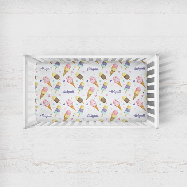 A white wooden baby crib with a custom printed mattress protector featuring a colorful ice cream cone pattern and the name 'Abigail' repeated in purple script, set against a white wood-paneled background.