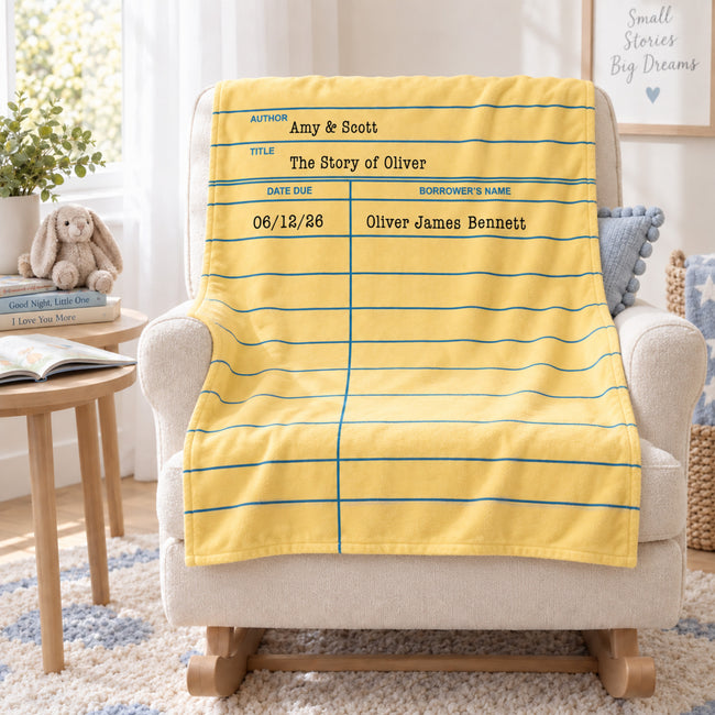 A yellow library card blanket with blue horizontal lines and printed text fields, draped over a cream armchair in a bright, styled room with a side table, rabbit toy, and framed wall art.
