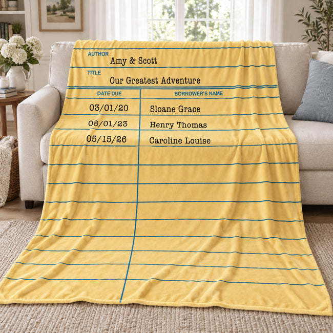 A yellow fleece library card blanket with a lined notebook paper design and printed text fields, draped over a beige sofa in a living room setting.