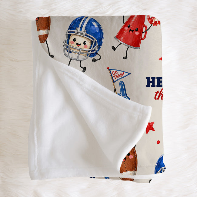 A folded baby blanket with a white plush reverse side and a printed front featuring cartoon football elements, including a blue helmet, red cheerleader hat, and football, on a cream background.