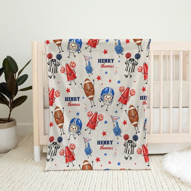 A personalized baby blanket with a red, white, and blue football theme, draped over a light wood crib in a nursery setting.