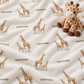 A close-up of a plush blanket or throw with an allover giraffe and text pattern. The design features brown giraffes and the name 'emerson' printed on a soft off-white background.