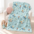A personalized blanket with an underwater mermaid theme, draped over a cream-colored armchair. The blanket features illustrations of mermaids, dolphins, fish, and the custom name 'Anna Catherine' printed in gold.