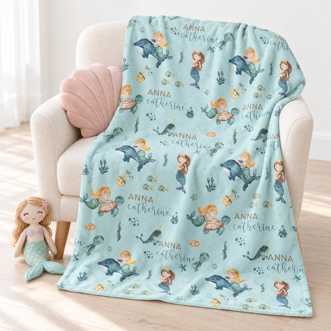 A personalized blanket with an underwater mermaid theme, draped over a cream-colored armchair. The blanket features illustrations of mermaids, dolphins, fish, and the custom name 'Anna Catherine' printed in gold.