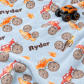Close-up of the Monster Trucks blanket personalized with Ryder, showing watercolor trucks on pale blue with an orange toy truck resting on top.