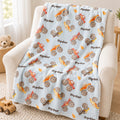 Personalized Ryder monster truck blanket draped over an ivory armchair, with colorful watercolor trucks and flames on pale blue.