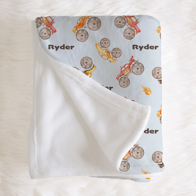 Folded Monster Trucks blanket with Ryder lettering and watercolor trucks, with a corner turned back to reveal the white reverse.