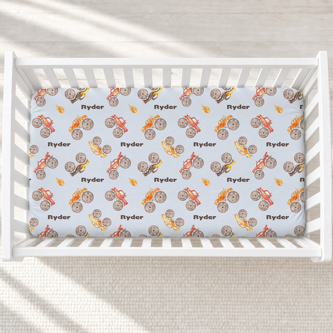 Monster Trucks personalized crib sheet with Ryder lettering and colorful watercolor trucks on pale blue, fitted in a white crib.