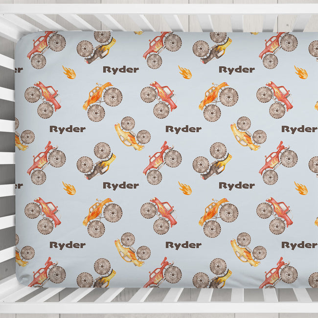 Close-up of the Monster Trucks crib sheet showing watercolor trucks, oversized tires, orange flames, and brown Ryder lettering.