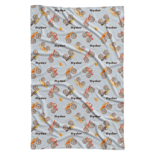 Full view of the Monster Trucks personalized swaddle blanket with brown Ryder lettering, watercolor trucks, and orange flame accents.