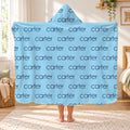 A person standing in a bedroom holding up a hooded blanket with a light blue background and a repeating dark blue 'carter' text pattern. The room has a window, wooden furniture, and a woven rug.