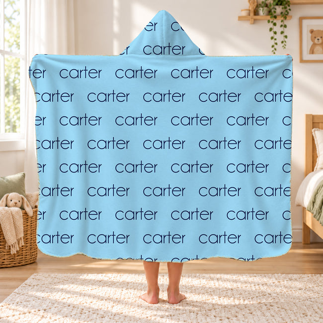 A person standing in a bedroom holding up a hooded blanket with a light blue background and a repeating dark blue 'carter' text pattern. The room has a window, wooden furniture, and a woven rug.