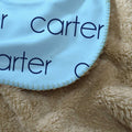A close-up of a light blue fabric item with dark navy text reading 'carter' laid over a beige plush textured blanket.