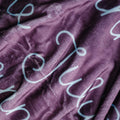 A close-up of a plush, textured fabric in a dusty purple color, printed with a repeating pattern of white cursive script letters.