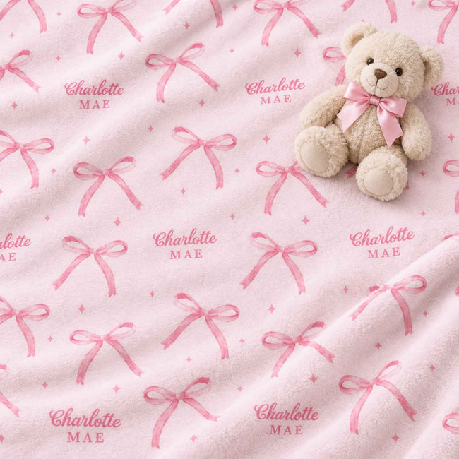 A plush light pink blanket with an all-over repeating pattern of pink bows and the custom text 'Charlotte MAE' in pink, styled with a small beige teddy bear wearing a matching pink bow on a soft white background.