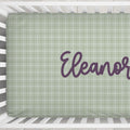 A fitted crib sheet with a sage green and white plaid pattern, personalized with the name 'Eleanor' in purple script, shown inside a white wooden crib.