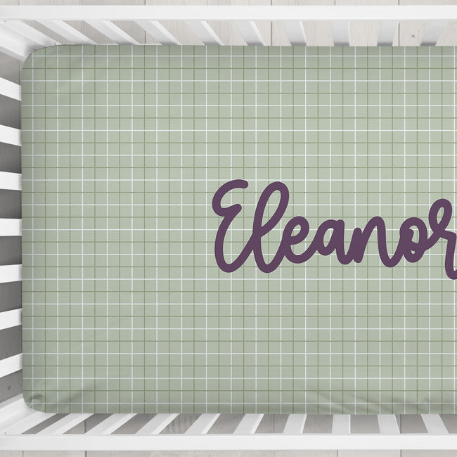 A fitted crib sheet with a sage green and white plaid pattern, personalized with the name 'Eleanor' in purple script, shown inside a white wooden crib.