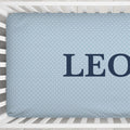 A light blue baby crib mattress cover with a white polka dot pattern and the name 'LEO' printed in navy blue block letters, shown inside a white wooden crib.