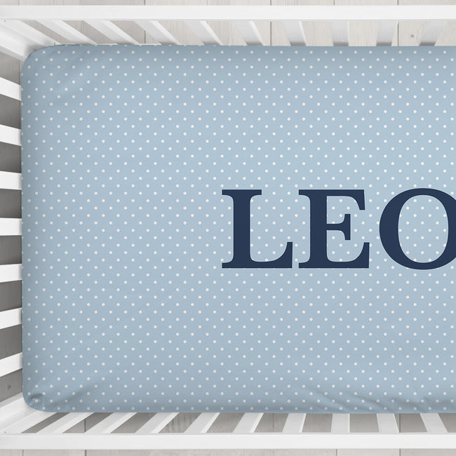 A light blue baby crib mattress cover with a white polka dot pattern and the name 'LEO' printed in navy blue block letters, shown inside a white wooden crib.