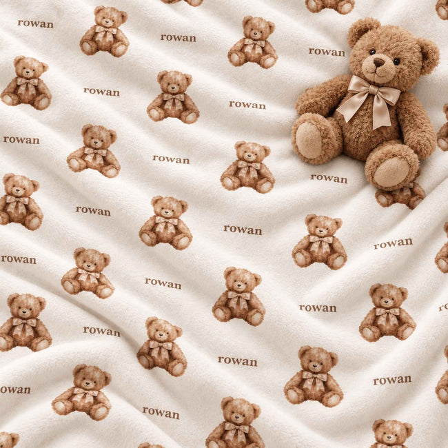 A close-up of a soft cream-colored fabric with an allover print of brown teddy bears and the name 'rowan' repeated in small text, with a plush teddy bear placed on the upper right.