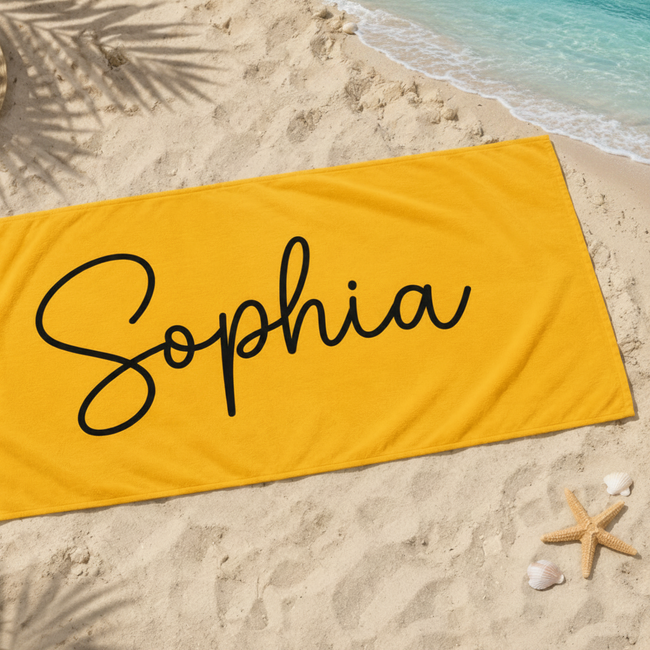 A bright yellow beach towel with the name 'Sophia' written in black cursive script, laid flat on a sandy beach with a straw hat, palm leaves, shells, and a starfish nearby.