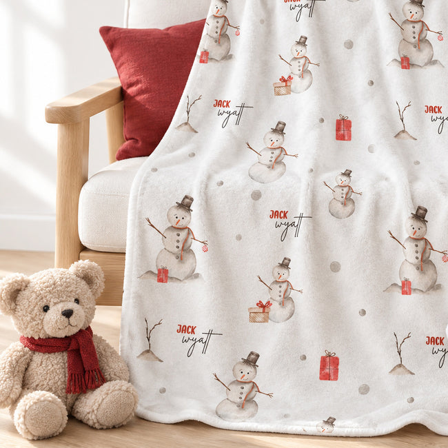 A white fleece throw blanket with a repeating snowman print and the name 'Jack Wyatt' is draped over a wooden chair next to a teddy bear wearing a red scarf.