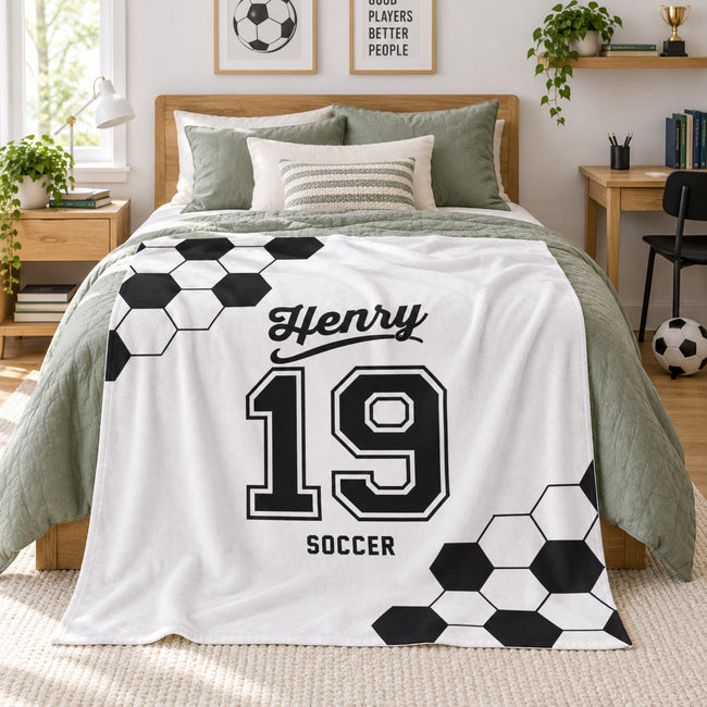 A personalized soccer-themed throw blanket draped over a bed in a modern, sunlit bedroom. The blanket is white with black soccer hexagon patterns and the text 'Henry 19 SOCCER'.
