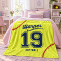A bright yellow personalized blanket styled on a bed in a pink-themed bedroom. The blanket features navy blue text reading 'Harper' and 'SOFTBALL' with the number '19' in the center, and red baseball stitching details.