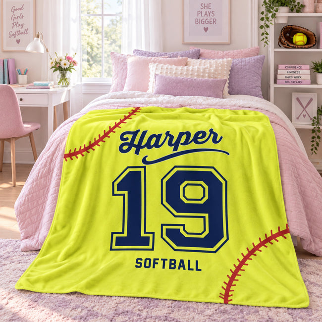 A bright yellow personalized blanket styled on a bed in a pink-themed bedroom. The blanket features navy blue text reading 'Harper' and 'SOFTBALL' with the number '19' in the center, and red baseball stitching details.