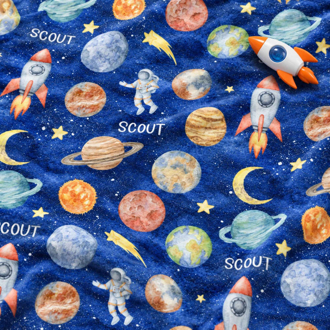 A close-up of a blue fabric with a colorful space-themed print featuring planets, stars, rockets, astronauts, and crescent moons, with the word 'SCOUT' repeated across the design.