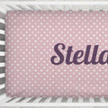 A pink fitted crib sheet with a white star pattern and the name 'Stella' printed in dark purple script, shown inside a white crib.