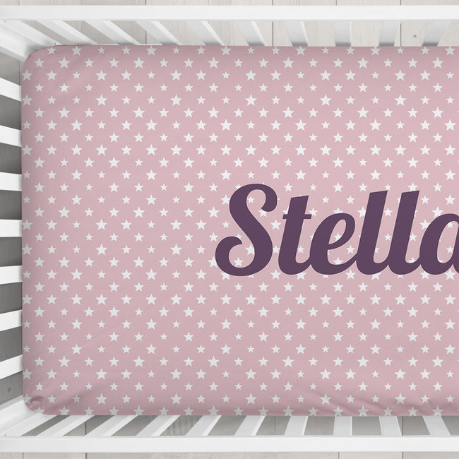 A pink fitted crib sheet with a white star pattern and the name 'Stella' printed in dark purple script, shown inside a white crib.